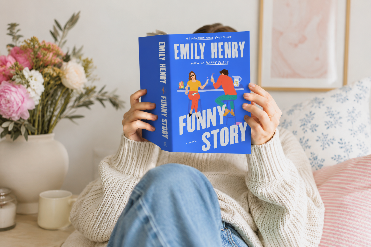 A person in a cozy cream knit sweater relaxes indoors while holding a copy of Funny Story by Emily Henry directly in front of their face, hiding their identity. The bright blue book cover stands out against the soft, neutral-toned room, which features a floral arrangement, a coffee mug, decorative pillows, and framed wall art, creating a warm, inviting reading atmosphere.